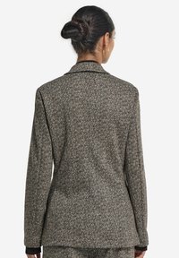 Kvinna med mörkt hår i en knut bär en kavaj i brun och beige fiskbensmönster, visad bakifrån mot en enfärgad vit bakgrund.