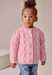 Cardigan rosa lavorato a maglia con motivo a trecce, bottoni in legno e polsini a coste, abbinato a jeans blu. Tessuto strutturato con screziature colorate.