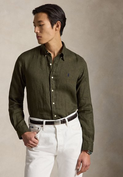 Egy férfi olívazöld gombos inget és fehér nadrágot visel fekete övvel, bézs háttér előtt áll, balra néz.