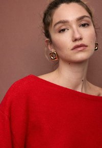 Roter, übergroßer Strickpullover mit weitem Ausschnitt und weicher Textur. Mit großen quadratischen goldenen Ohrringen und neutralem Hintergrund.