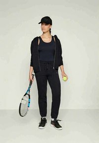 Γυναίκα με μαύρη αθλητική ενδυμασία κρατά ρακέτα τένις με το δεξί χέρι και μπάλα τένις με το αριστερό, στέκεται μπροστά σε απλό φόντο.