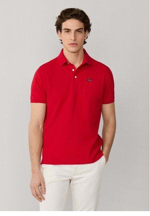 Hombre con una camiseta polo de manga corta roja con botones blancos y pantalones blancos, de pie con una mano en el bolsillo contra un fondo liso.