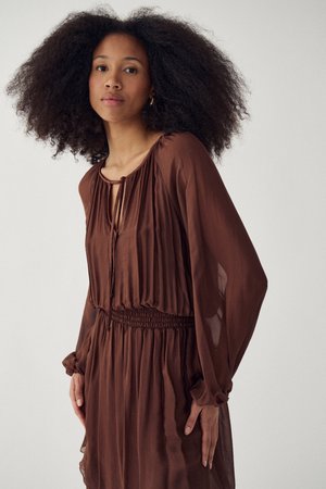 Kvinde med naturligt krøllet hår iført en brun kjole med lange ærmer, markeret talje og nøglehulshalsudskæring, stående mod en ensartet lys baggrund.
