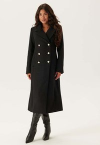 Cappotto doppiopetto in lana nera con grandi bottoni bianchi, colletto a scollo a V e due tasche frontali, abbinato a stivali neri al ginocchio.