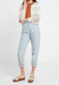 Cardigan bianco all'uncinetto, top fitted color ruggine, jeans azzurri con risvolto, e sandali bianchi su zeppa di legno.