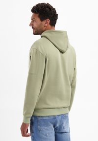 Capuchontrui in zachte olijfgroene stof, met een klassieke pasvorm, geribbelde manchetten en een kangoeroezak. Achteraanzicht toont een eenvoudig ontwerp.