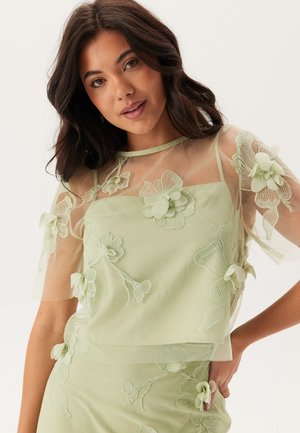 Haut vert clair en tissu transparent, avec broderies florales et manches courtes. Présente un design superposé avec des fleurs en tissu et un col rond.
