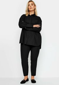 Camisa negra de botones con mangas largas, de corte amplio, y un bolsillo en el pecho, combinada con pantalones ajustados negros y mocasines negros.