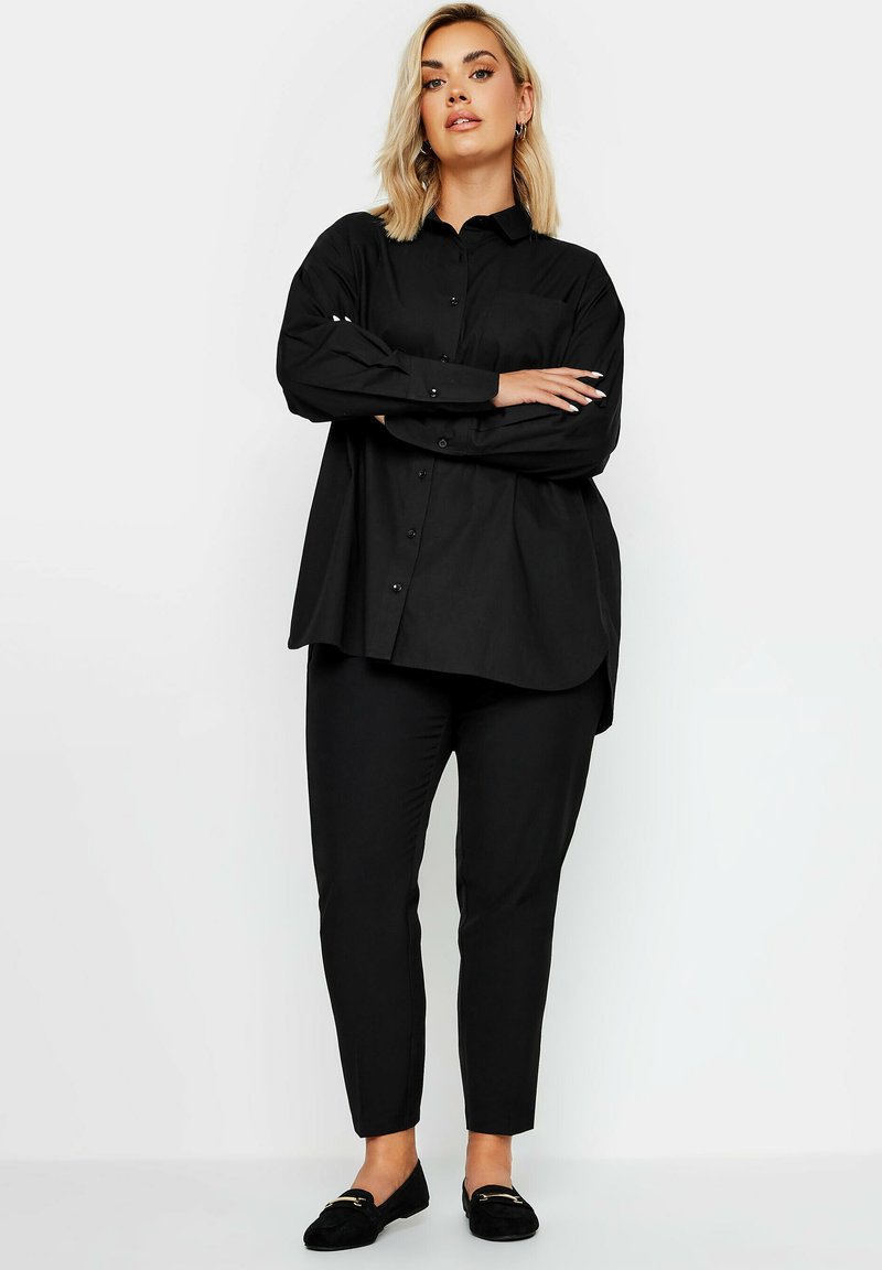 Camisa negra de botones con mangas largas, de corte amplio, y un bolsillo en el pecho, combinada con pantalones ajustados negros y mocasines negros.