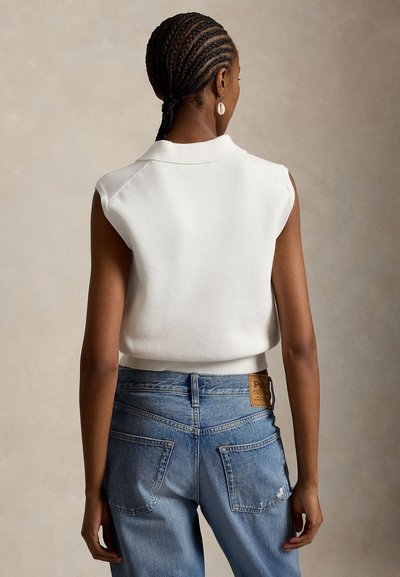 Moteris su pintais plaukais, apsirengusi baltą rankovių megztinį ir mėlynas džinsines kelnes, stovinti atsukusi nugarą prieš vienspalvį foną.