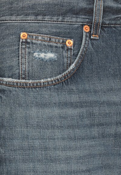 Jeans in denim con un colore blu, con una tasca in evidenza decorata con rivetti in rame e un'area strappata e consumata vicino alla cucitura della tasca.