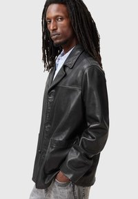 Schwarze Lederjacke mit Reverskragen, zwei Vorderaschen und einem knopfartigen Verschluss. Das Modell trägt ein hellblaues Hemd darunter.
