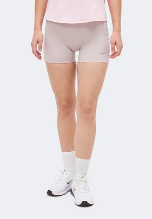 Άτομο που στέκεται φορώντας ανοιχτόροζ σορτς, λευκές κάλτσες μέχρι τον αστράγαλο, λευκά αθλητικά παπούτσια Nike και ανοιχτόροζ μπλούζα, με μονόχρωμο φόντο.