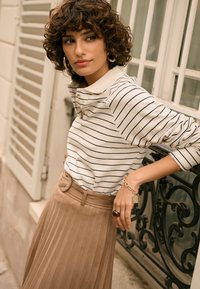 Maglia a righe a maniche lunghe in beige e blu navy, abbinata a una gonna plissettata color tan. Gioielli dorati di rilievo e cintura con bottoni completano il look.