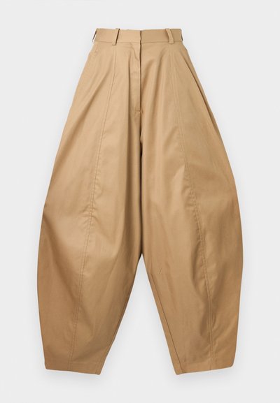 Kõrge vöökoht, laiad beezid püksid voldikute, vöökaarte ja kitsenevate pahkluude abil, esitlemine valge tausta ees.