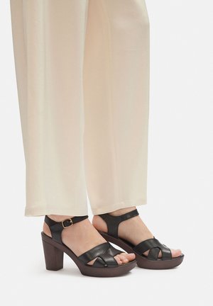 Czarne sandały na platformie z paskami krzyżującymi się i klamrą przy kostce, z brązowym, blokowym obcasem, zestawione z jasnobeżowymi szerokimi spodniami.