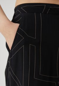 Černá tkanina s texturovaným povrchem má bílou přerušovanou nit v geometrických vzorech. Na levé straně je viditelná kapsa.