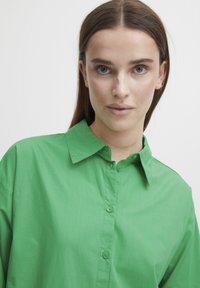 Green collared shirt made of lightweight fabric, featuring a button-up front, and long sleeves with a smooth texture and subtle sheen.