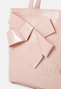 Sac fourre-tout rose brillant avec un grand nœud plat. L'avant affiche "Ted Baker LONDON" en texte doré en relief. Texture lisse, forme douce.