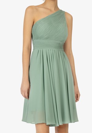 Kobieta ubrana w jasnozieloną, plisowaną sukienkę na jedno ramię sięgającą do kolan, z dopasowaną talią i zwiewną spódnicą.