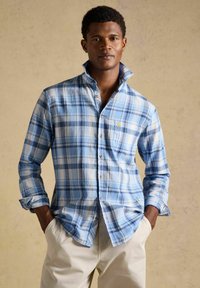 Nuori mies, joka pukeutunut siniseen ruudulliseen kaulukselliseen nappipaitaan ja beigenvärisiin housuihin, seisoo kädet taskuissa neutraalia taustaa vasten.