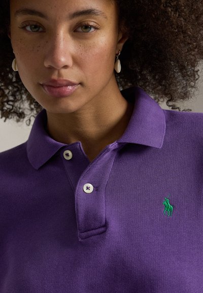 Μωβ πόλο πουκάμισο κατασκευασμένο από υφασμένο ύφασμα, με πατιλέτα τριών κουμπιών και πράσινο κεντημένο λογότυπο. Λευκά κουμπιά με μωβ ραφές.