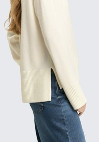 Maglione color beige chiaro in morbido tessuto lavorato a maglia, con polsini a coste e spacco laterale all'orlo, abbinato a jeans di denim blu.