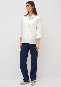 Lichtbeige overhemd met knopen en driekwart mouwen, van een gestructureerde stof, en marineblauwe rechte jeans. Casual schoeisel met groene accenten.
