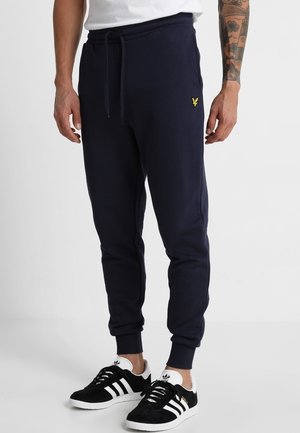 Mann trägt dunkelblaue Jogger mit einem gelben Logo am linken Oberschenkel und schwarzen Adidas-Sneakern mit weißen Streifen und Sohlen.