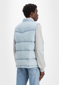 Gilet matelassé bleu clair avec un design en coussinets et un col haut. Assorti à une chemise à manches longues grise et un bas en denim clair.
