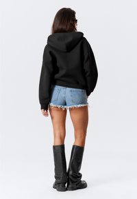 Svart oversized huvtröja, blå denimshorts med franskant och knähöga svarta stövlar, sedd bakifrån mot en enfärgad bakgrund.