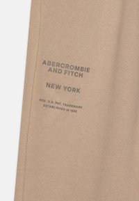 Beżowa tkanina bawełniana z nadrukowanym tekstem "ABERCROMBIE AND FITCH" oraz "NOWY JORK" w ciemnoszarym kolorze. Gładka tekstura bez dodatkowych wzorów ani akcentów.