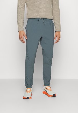 Personne portant un pantalon de jogging bleu-gris noué à la taille, un pull texturé beige et des baskets blanches avec des semelles et des lacets orange.
