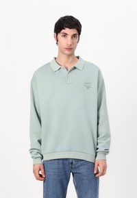 Light green long-sleeve polo shirt with a collar, featuring a subtle logo on the chest, paired with blue denim jeans.
