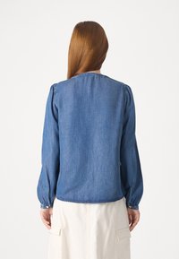 Femme aux longs cheveux roux et lisses, vue de dos, portant une chemise en denim bleu à manches longues et une jupe blanche avec des poches.