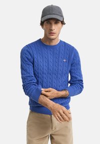 Blauwe kabelgebreide trui met een ronde halslijn, geribde manchetten en zoom. Draagt een grijze baseballpet. Beige broek zichtbaar.