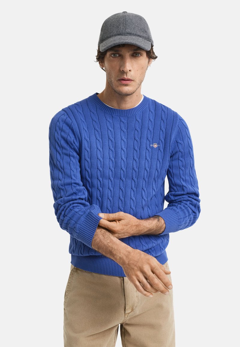 Blauwe kabelgebreide trui met een ronde halslijn, geribde manchetten en zoom. Draagt een grijze baseballpet. Beige broek zichtbaar.