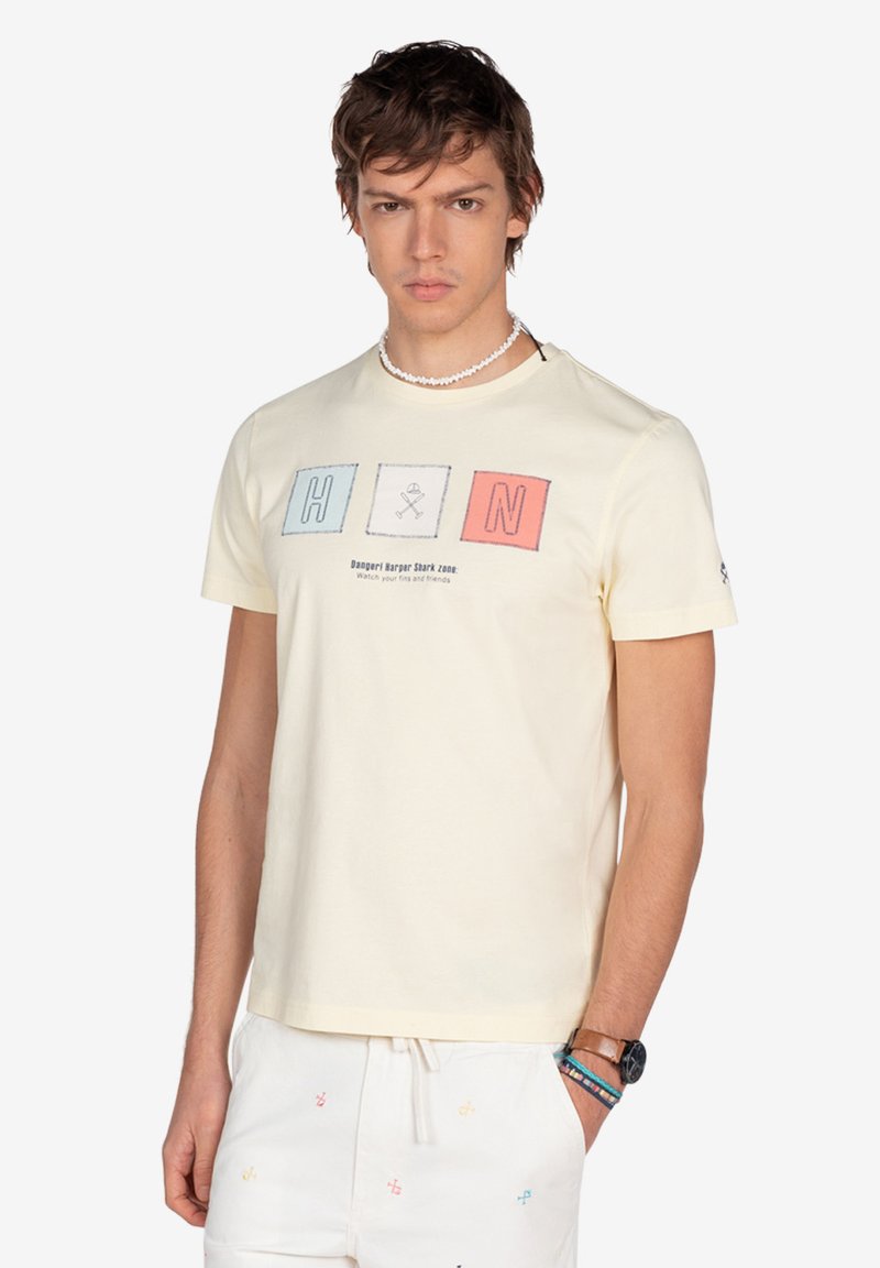 Camiseta de algodón de manga corta, de color amarillo claro, con letras en bloque "H" y "N" en azul y rojo, acompañadas de un gráfico y texto debajo de las letras.