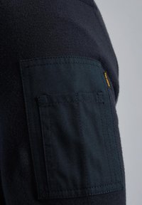 Pull marin avec un tricot texturé et une poche en tissu sombre sur la manche. Étiquette de logo jaune près de la couture de la poche.