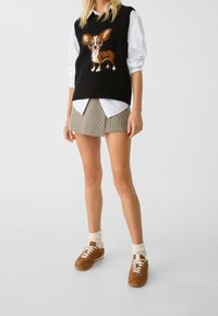 Gilet noir en tricot avec un motif de chien marron et blanc, superposé sur une chemise blanche, associé à une mini jupe à carreaux et des baskets marron.