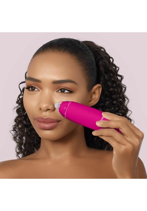 Femme aux cheveux bouclés utilise un appareil de nettoyage facial rose vif sur sa joue sur un fond violet pâle.