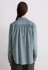Helles blaues langärmliges Hemd mit plissiertem Rückendesign, geknöpftem Kragen und weicher Textur, von hinten präsentiert.