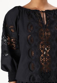 Blusa nera con intricati ricami in pizzo, scollatura a girocollo e maniche a sbuffo con polsini elasticizzati; nastro decorativo nella parte anteriore.