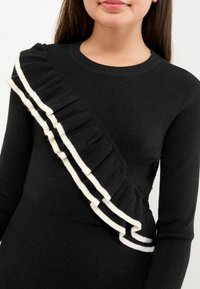 Sort sweater med et asymmetrisk design, der har en ruflet kant i creme, hvilket indikerer tekstur og bevægelse. Ribbet halsudskæring og manchetter.