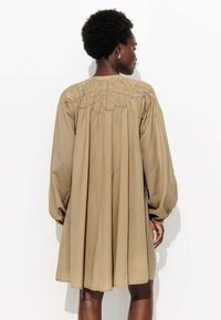 Robe longue beige avec un détail de corsage froncé, de longues manches bouffantes et une silhouette fluide. Tissu doux et texturé avec une coupe décontractée.