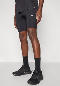 Μαύρα αθλητικά σορτς με λεία υφή και εφαρμοστό σχέδιο, συνδυασμένα με μαύρα αθλητικά παπούτσια και κάλτσες. Περιλαμβάνει λεπτομέρεια με λογότυπο.