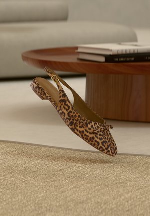 Leopardmønstret slingback flad sko med lille sløjfedetalje svævende over beige flettet tæppe nær rundt træ sofabord med bøger.