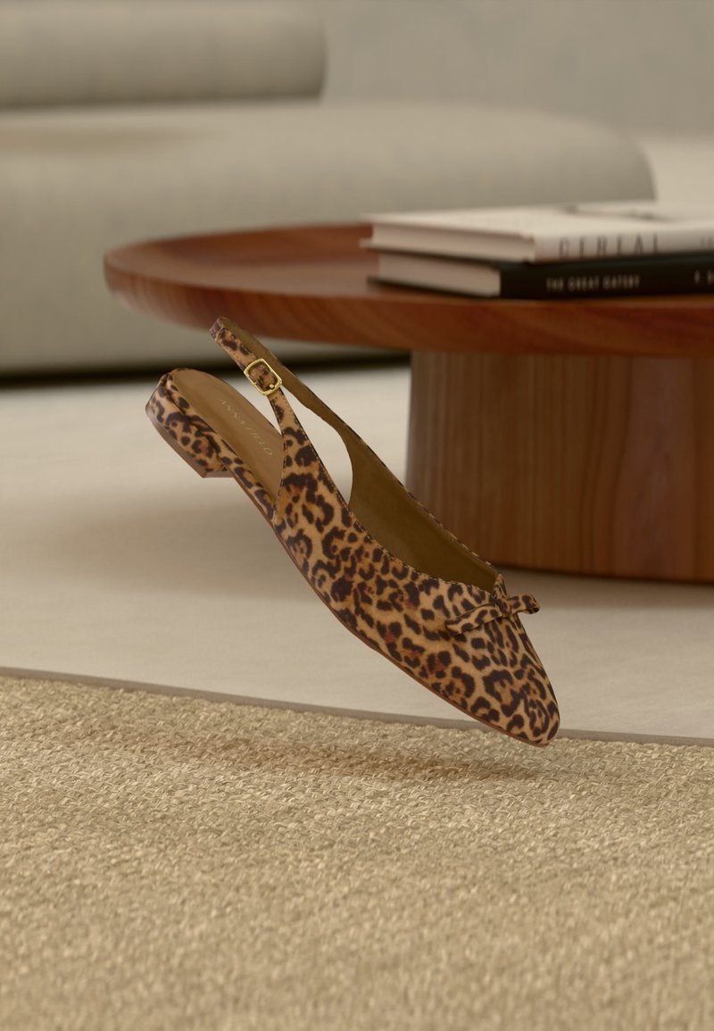 Chaussure plate slingback à imprimé léopard avec un petit nœud flottant au-dessus d'un tapis beige tissé, près d'une table basse ronde en bois avec des livres.
