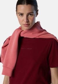 Camiseta marrón con el texto "NORTH SAILS", combinada con un sudadera rosa claro que cuelga sobre los hombros, con un tejido suave y un corte relajado.