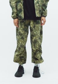 Persoon in loszittende groen en bruine camouflagebroek, witte sokken, zwarte sneakers en een bijpassende camouflagejas over een zwart shirt.
